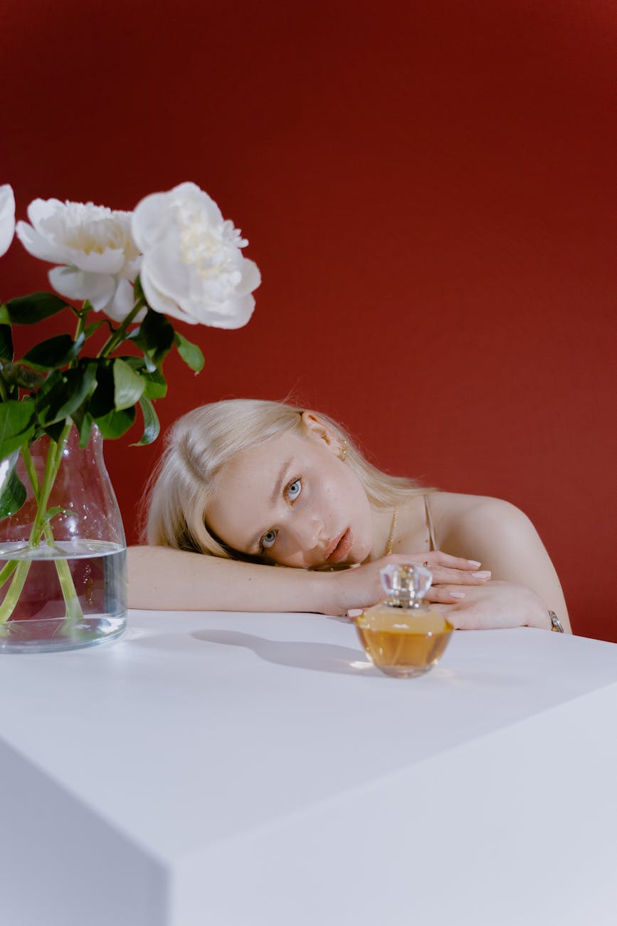 blonde girl leaning on white table with bottle of perfume and glass vase