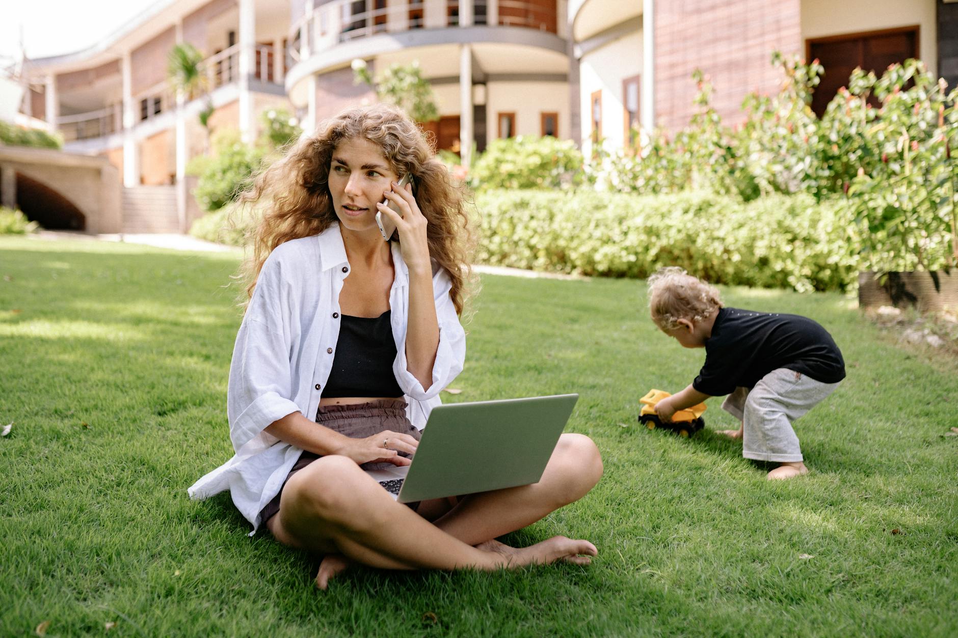 photo of woman using silver laptop with her child playing on grass field