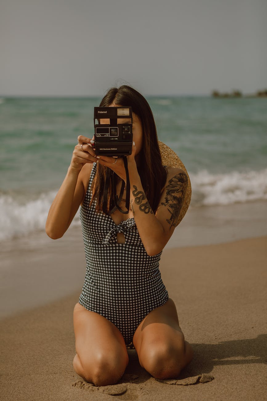 unrecognizable slender lady taking instant photos on vintage camera while resting on sandy sea coast