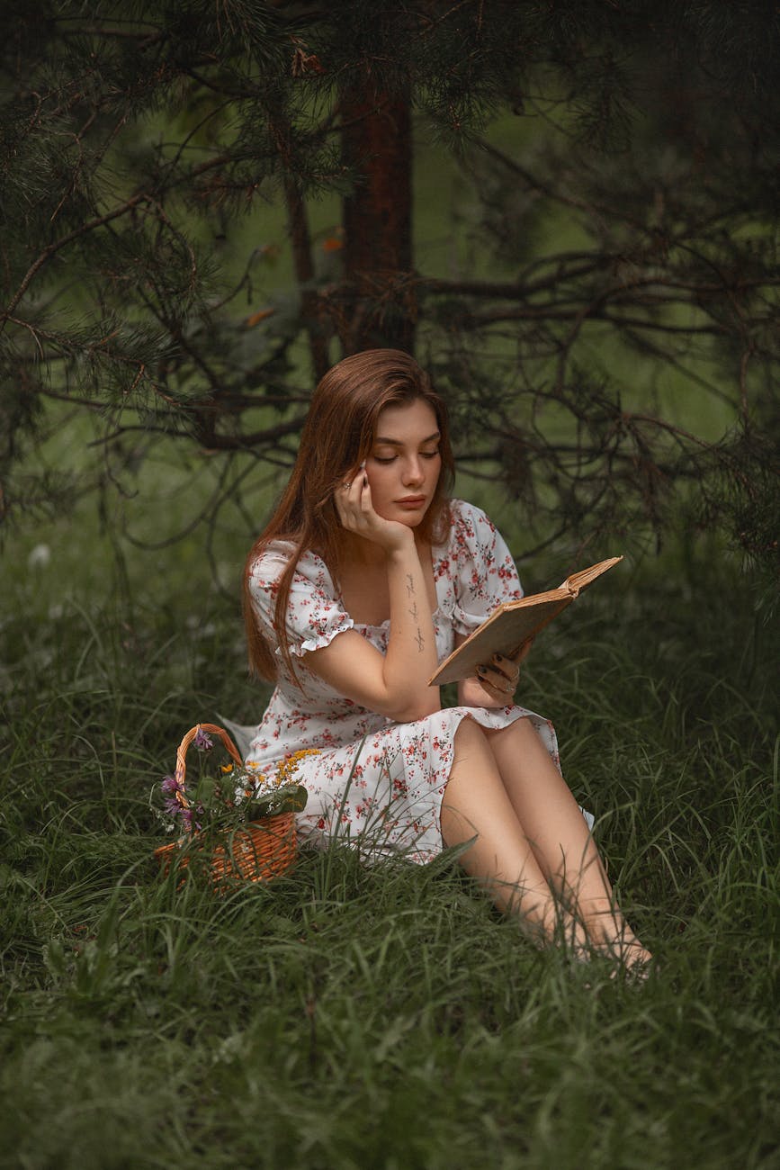 young woman in a dress sitting on the grass with a book