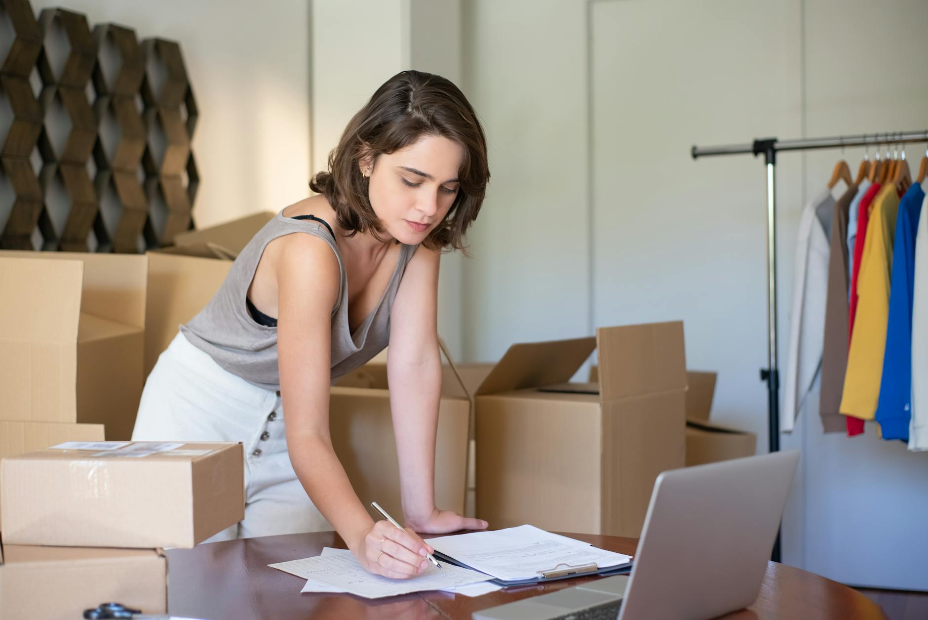 woman standing among cardboard boxes and writing on a piece of paper on a desk with a laptop